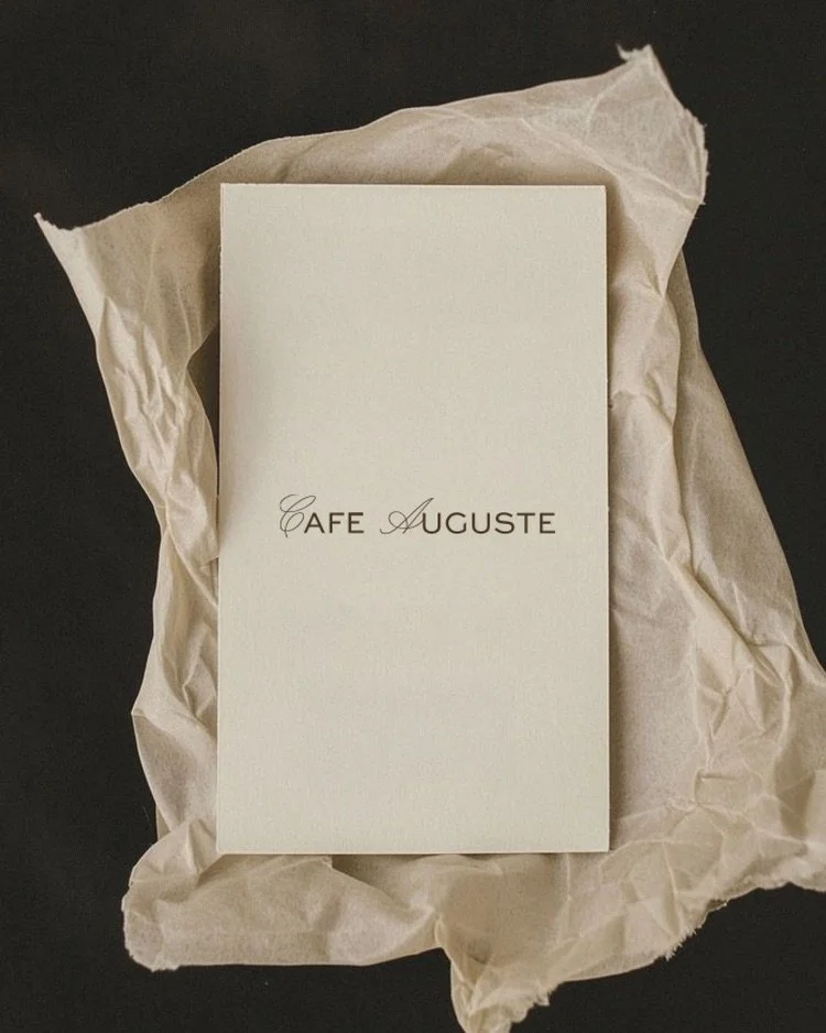 A white envelope with the words 'Café Auguste' printed in black in the center, resting on crumpled beige tissue paper against a dark background.