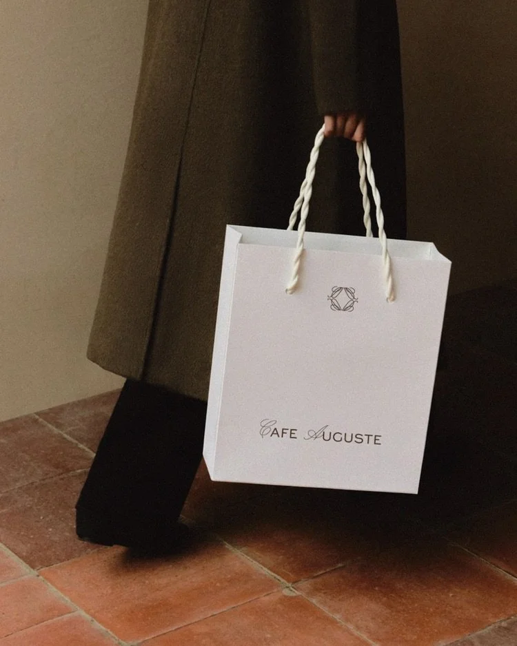 Person holding a white paper shopping bag with twisted rope handles, labeled 'Café Auguste,' next to a piece of furniture and tiled floor.