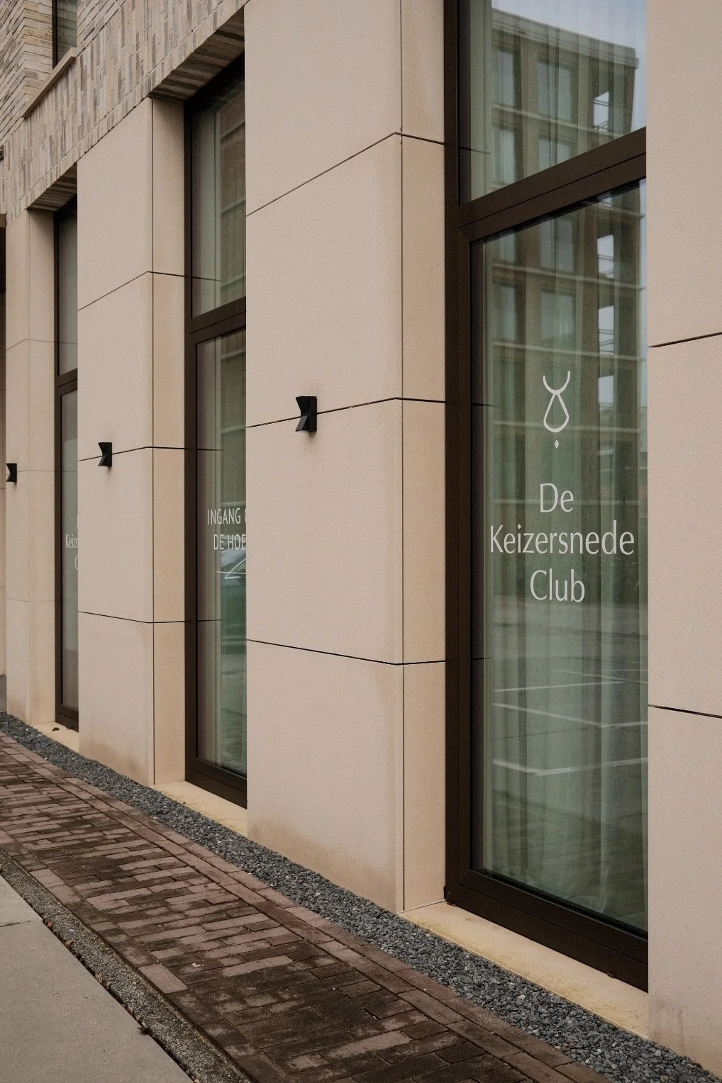 Exterior of a modern building with large glass windows and beige facade, with a sign reading "De Keizersnede Club" and an entrance sign in Dutch.