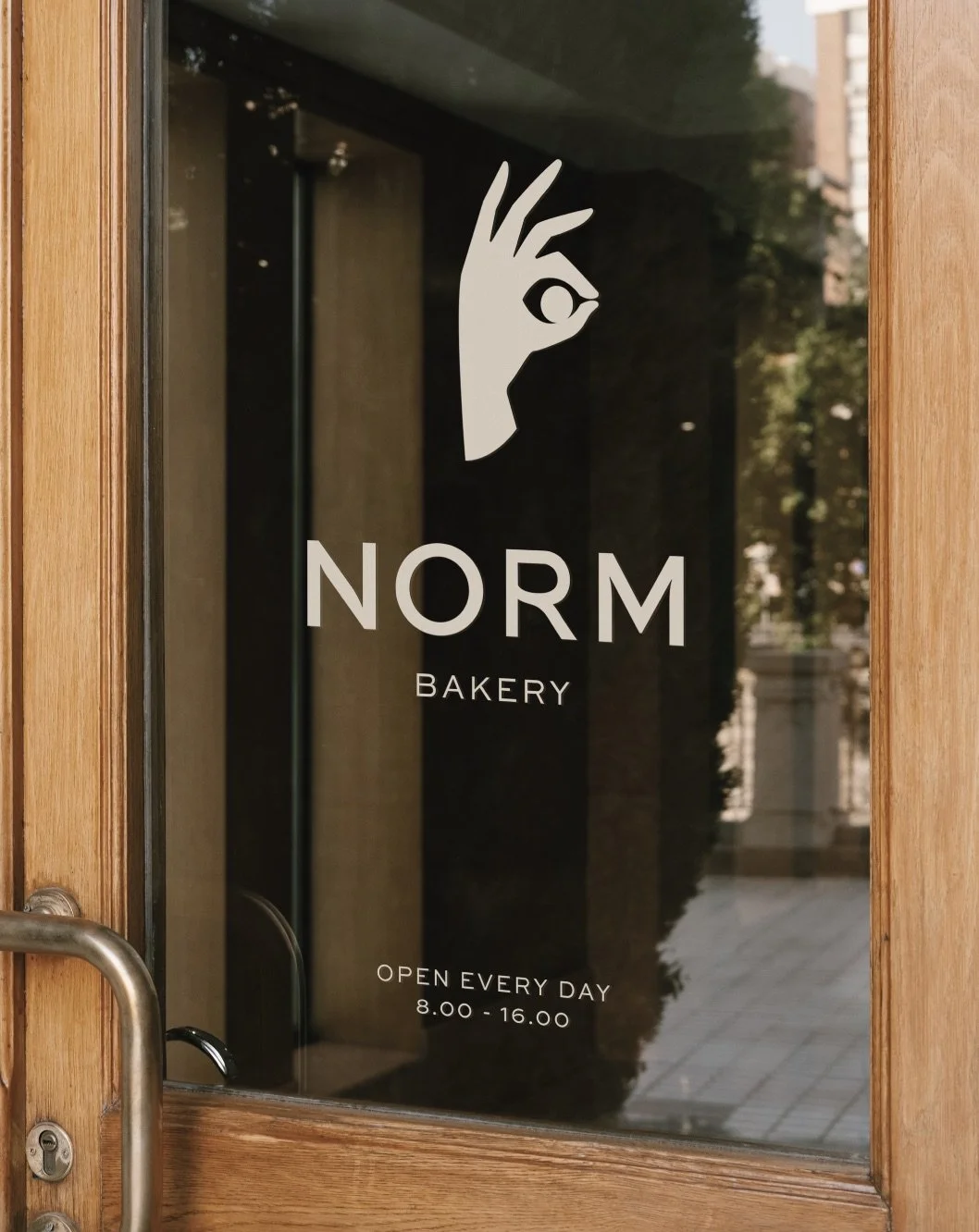 Glass door with a logo of a hand making an 'OK' gesture with an eye in the middle, the text 'NORM BAKERY' and opening hours 8:00-16:00, open every day.