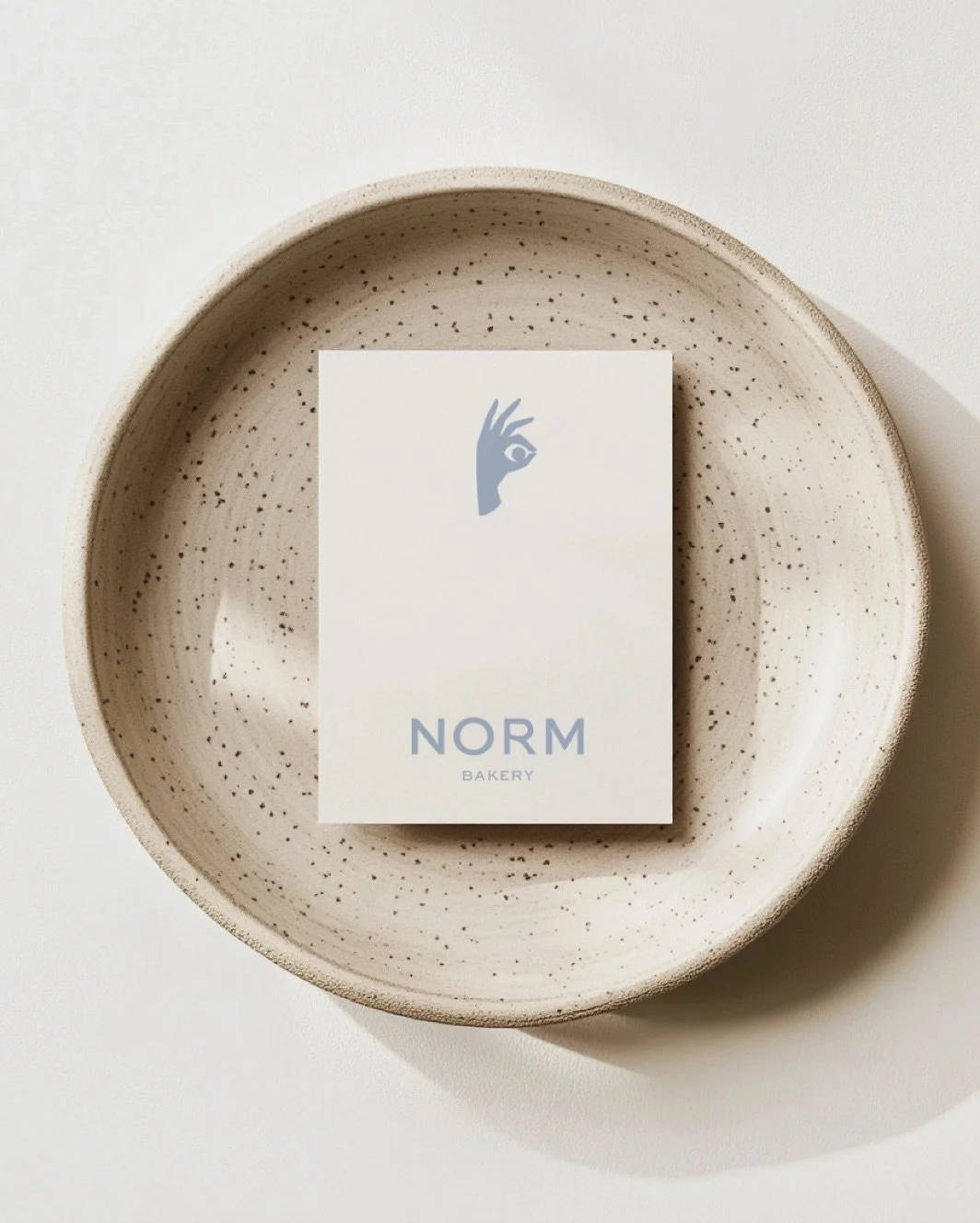 A speckled beige bowl with a business card inside that says "NORM Bakery" with an illustration of a hand forming an OK gesture.