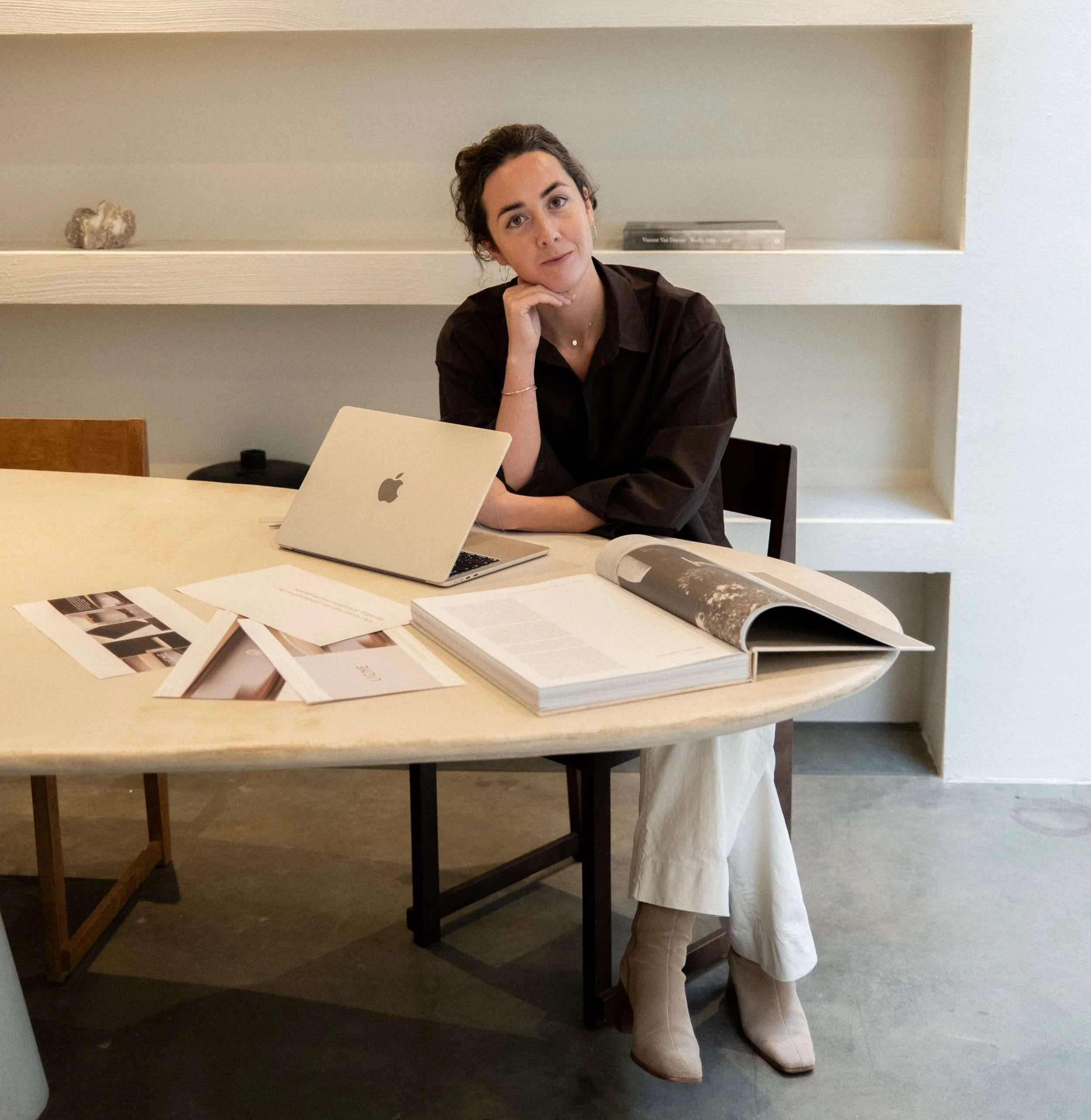 A woman walking in a beige creative studio, table in front of with laptop and paper designs