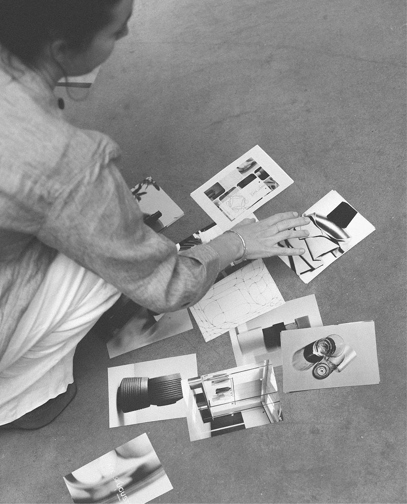 A person sitting on the floor with various photographs, sketches, and a small scale model of a building or object laid out in front of them.