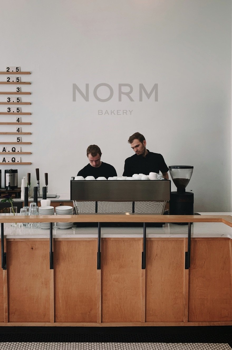 Two baristas work behind a counter at Norm Bakery, with a chalkboard displaying sizes on the left and coffee beans on the right.