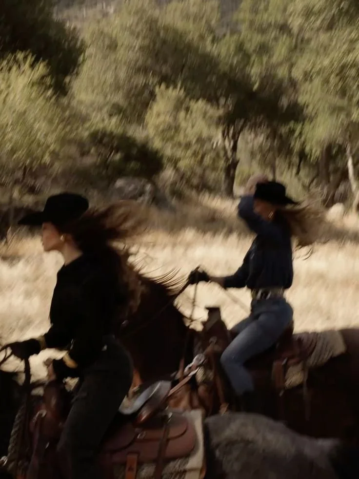 Two women riding horses outdoors at night, one in a black hat and jacket, the other in a black hat and jeans, with trees and dry grass in the background.