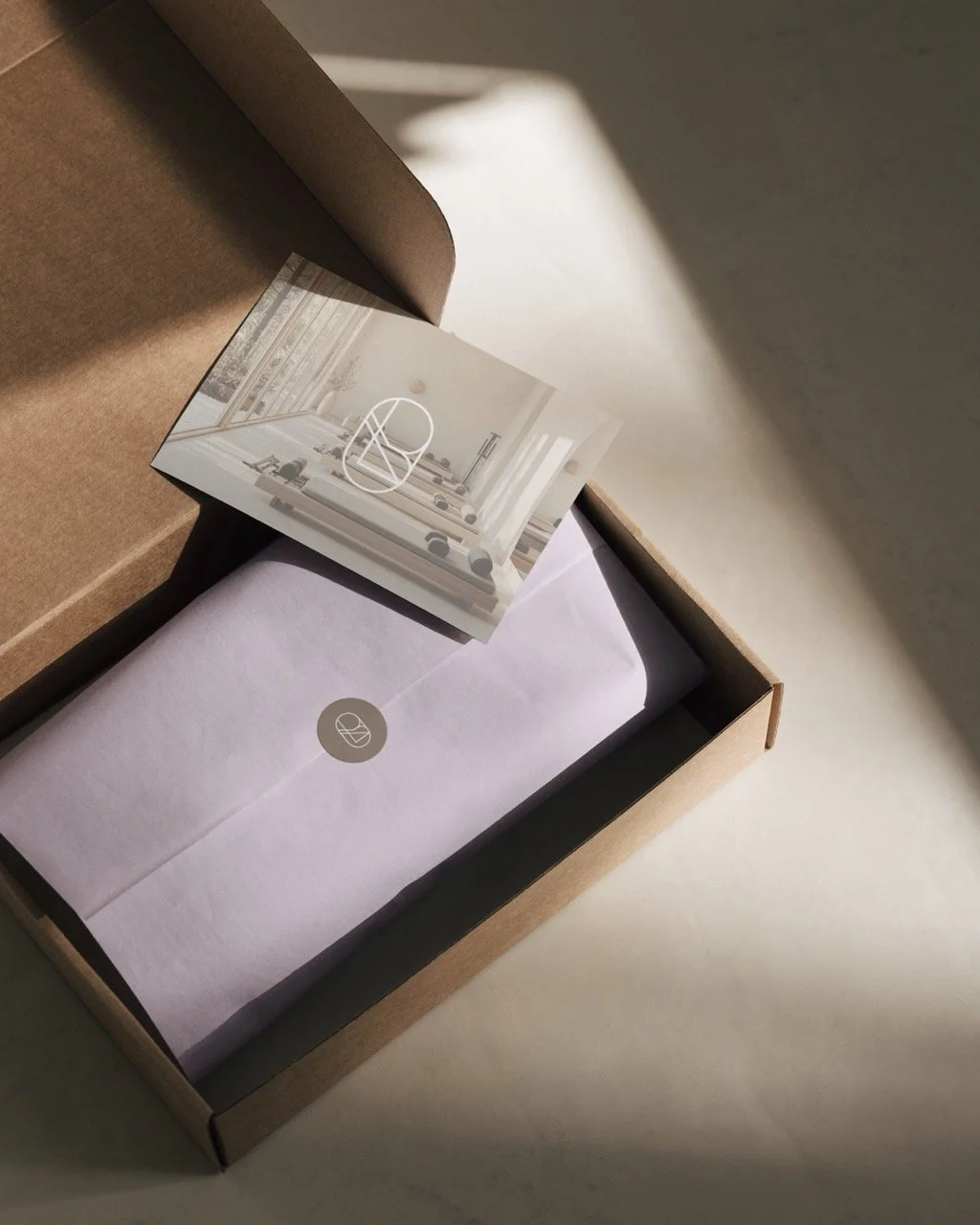 An open brown cardboard box containing a white envelope and a small card showing a modern, minimalist interior space with light-colored furniture and large windows.