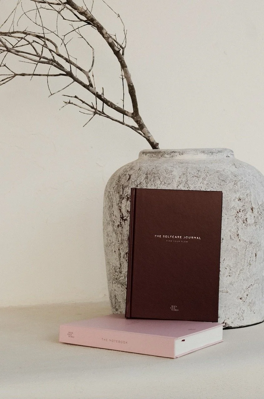 A large beige textured vase with a dry, leafless branch inside it. In front of the vase are two notebooks, one dark brown titled 'The Selfcare Journal' and one pink titled 'The Notebook'.