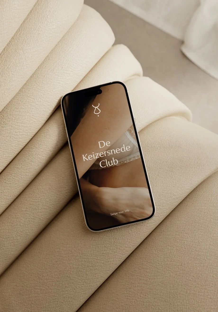 Smartphone resting on a beige upholstered chair with text on the screen that reads 'De Keizersnede Club'.
