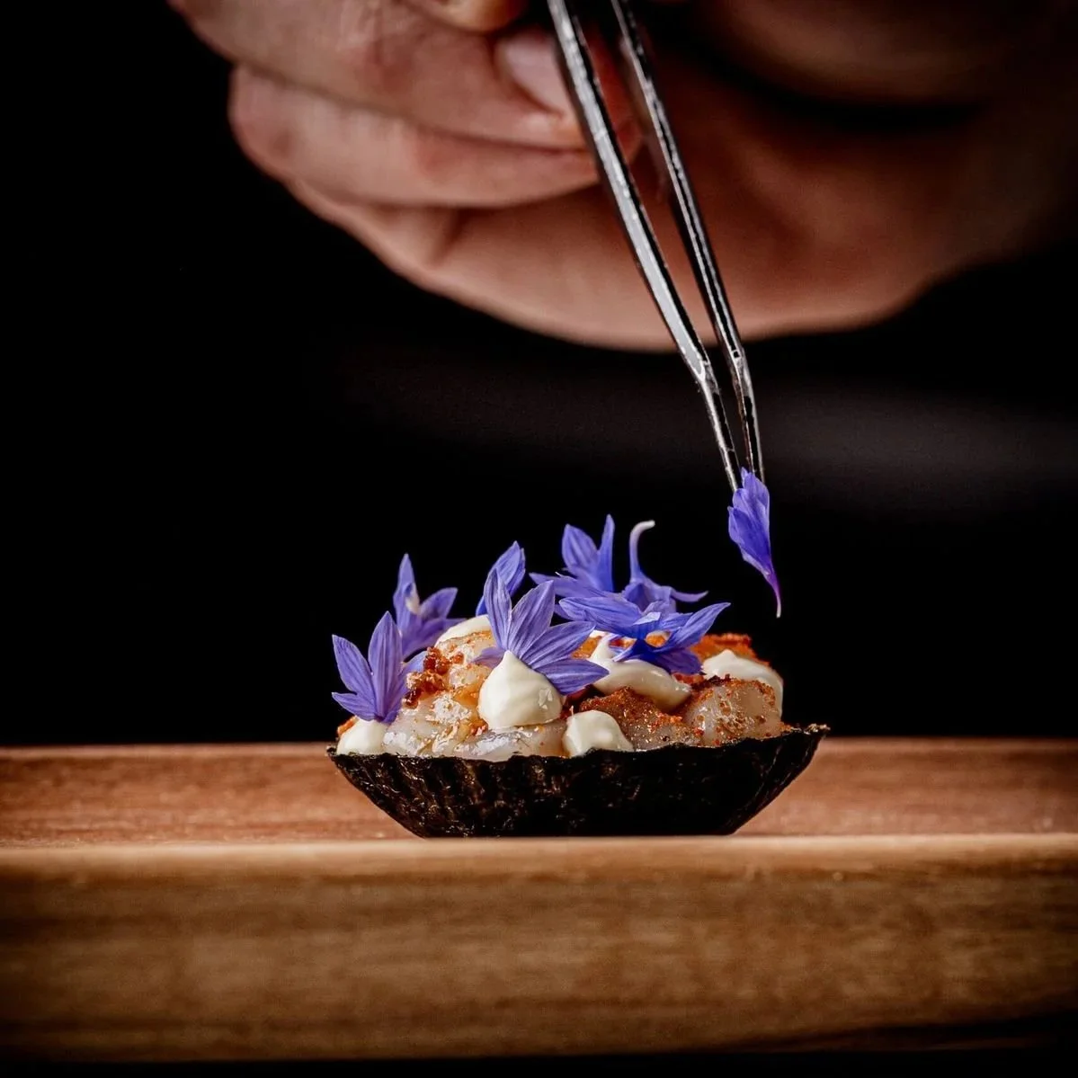 Close-up of a black bowl with sushi topped with white sauce and purple edible flowers, with a hand holding tweezers adding a purple flower, against a dark background.