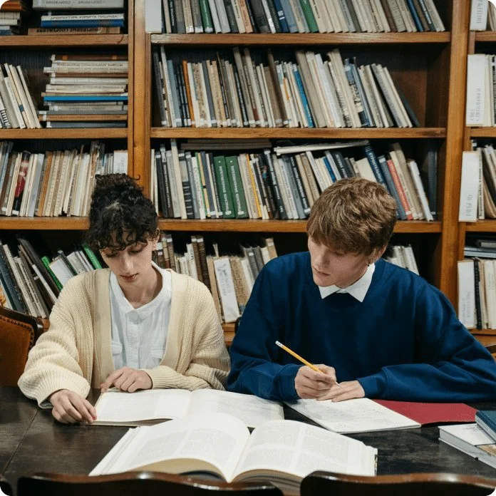 Two students studying together at a wooden table in a library with a large bookshelf filled with books behind them. One student is reading a book, the other is taking notes.