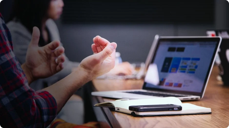 Person gesturing with hands during a meeting, laptop on the table showing a website or app, notebook and smartphone nearby, others in the background