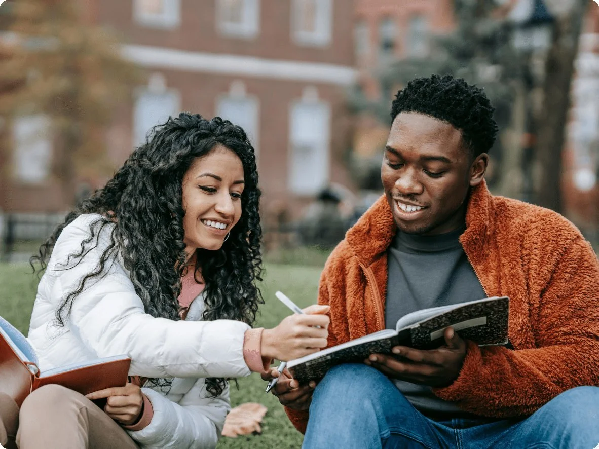A young woman and man sit on the grass in a park, smiling and looking at a notebook together, with the woman holding a pen.
