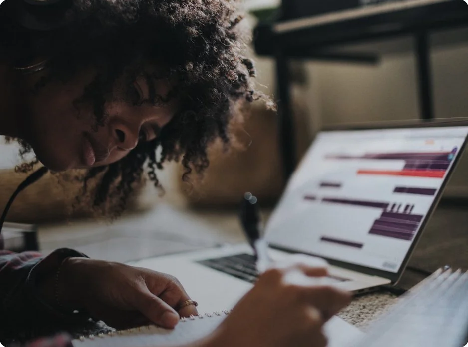 A woman with curly hair working on a graph on a laptop while writing in a notebook.