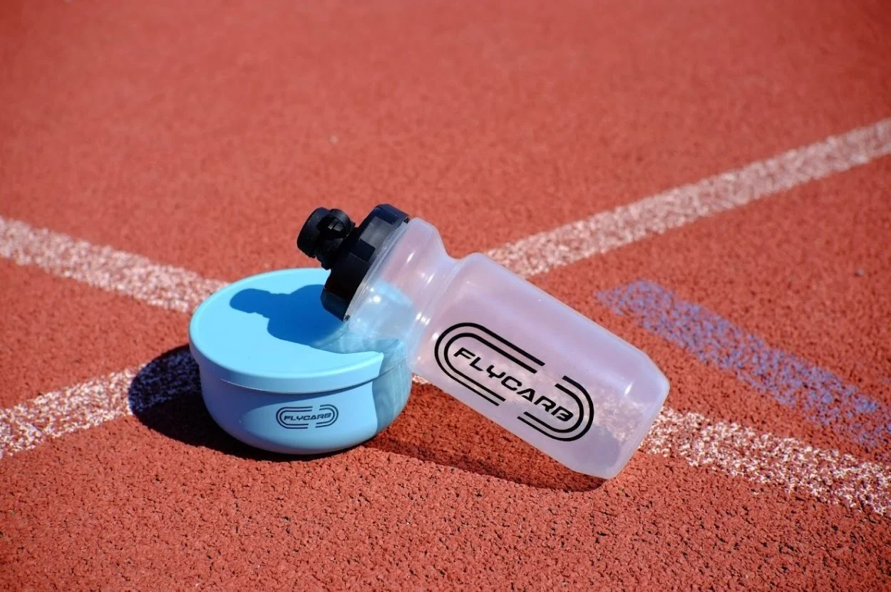 A Flycarb water bottle resting on a blue container on a red running track.