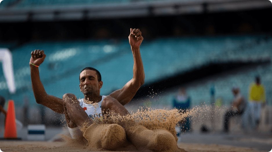 An athlete landing in a sandpit during a long jump event at an outdoor stadium with spectators in the background.