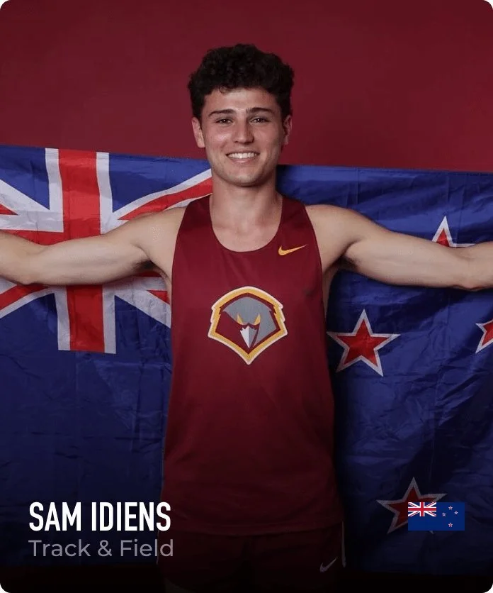 A young man with curly dark hair wearing a maroon athletic tank top with a logo, smiling, with the New Zealand flag in the background
