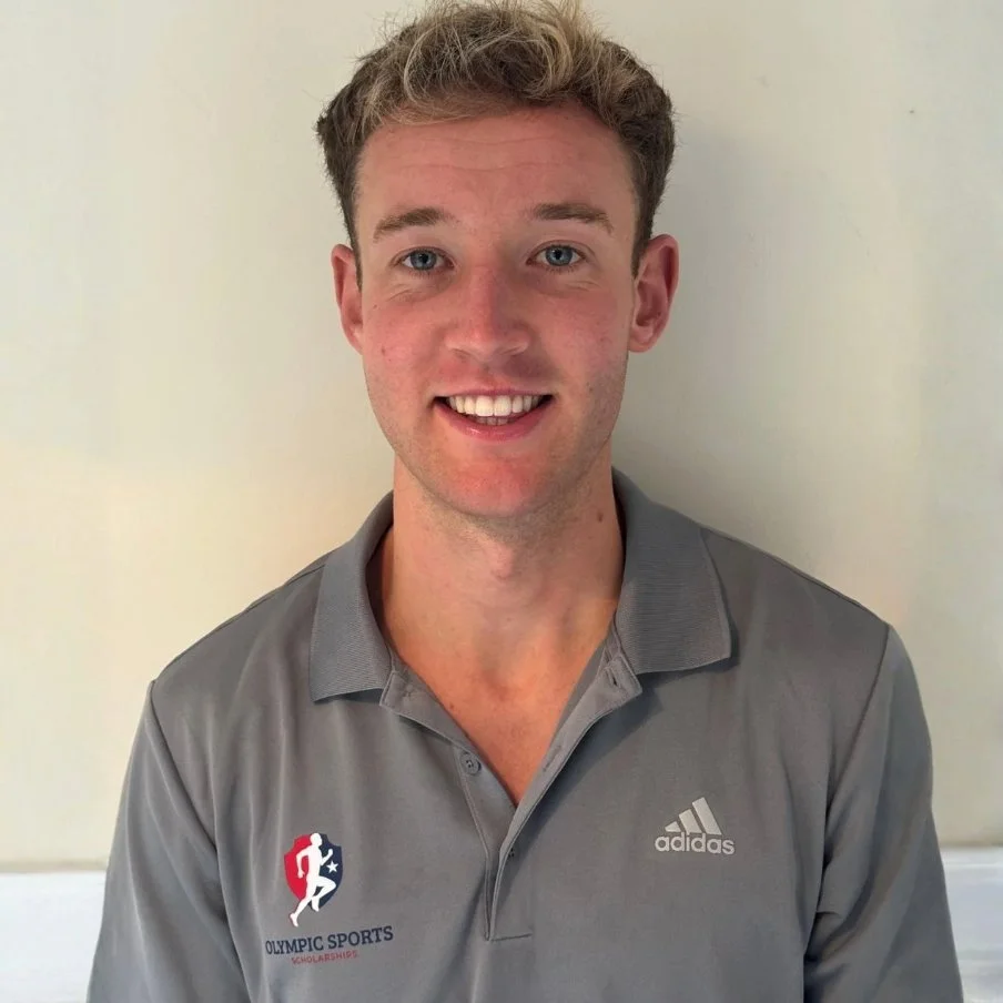 Young man with short, curly brown hair and blue eyes, smiling, wearing a gray Adidas polo shirt with 'Olympic Sports Scholarship' logo, standing against a plain white wall.