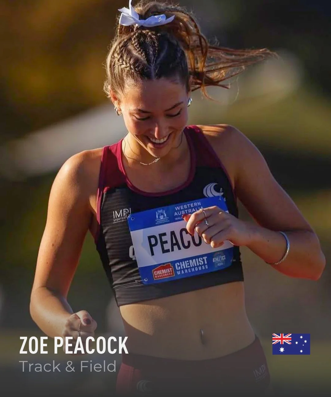 A woman with braided hair and a blue bow, wearing a red sports bra and black athletic gear, smiling and looking down while running outdoors during a track and field event, with a name tag that reads 'PEACOCK'.