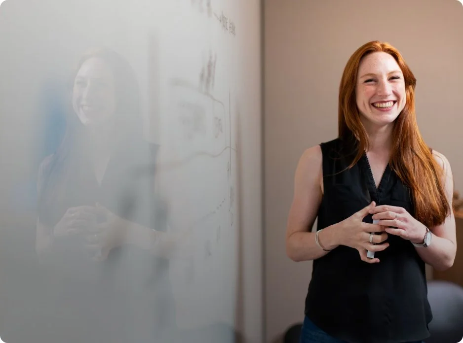 Red-haired woman smiling and standing next to a whiteboard in a classroom or office setting.