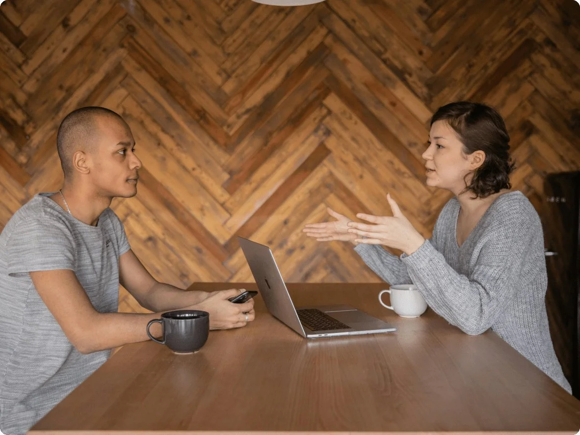 Two women having a serious conversation at a wooden table, with a laptop and coffee mugs in a room with wooden wall decor.