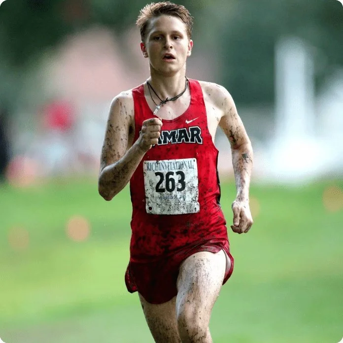 Young male cross-country runner covered in mud, wearing a red sleeveless jersey with 'GAMAR' printed on it, running on a grassy course during a race.