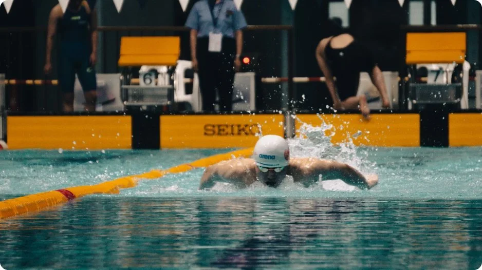 Swimmer in a swimming race doing a butterfly stroke at a pool, with officials and spectators in the background.