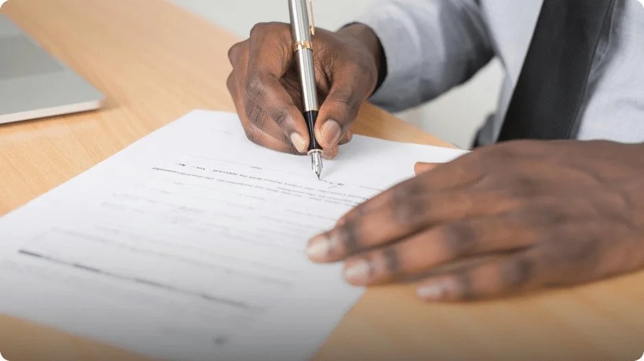 Person signing a document with a pen on a wooden desk