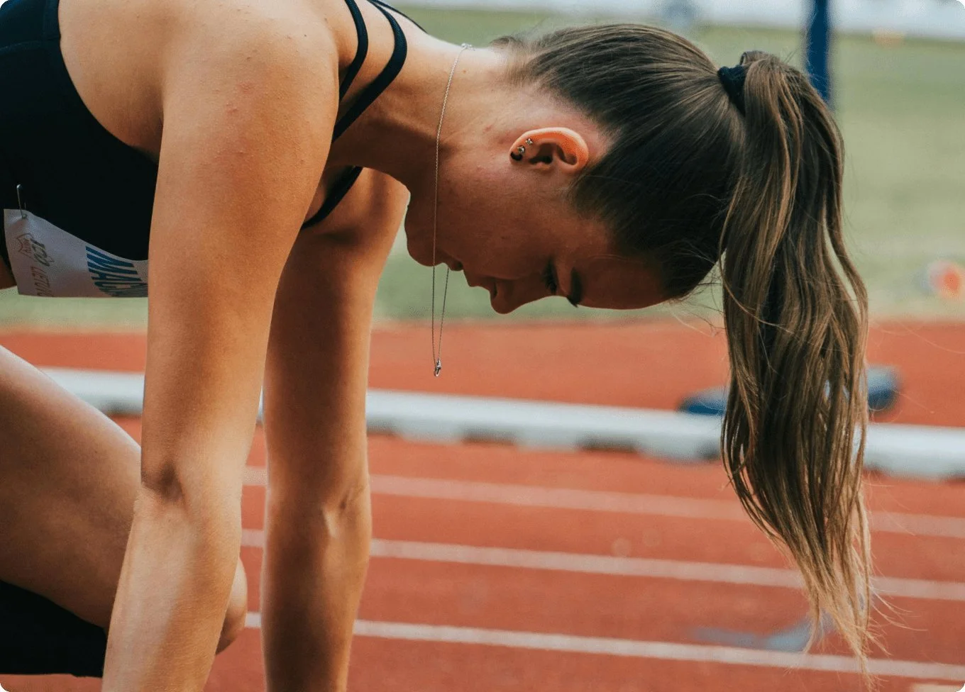 A female athlete on a running track, bent over with her head down, wearing a black sports top and a necklace, with her hair tied back.