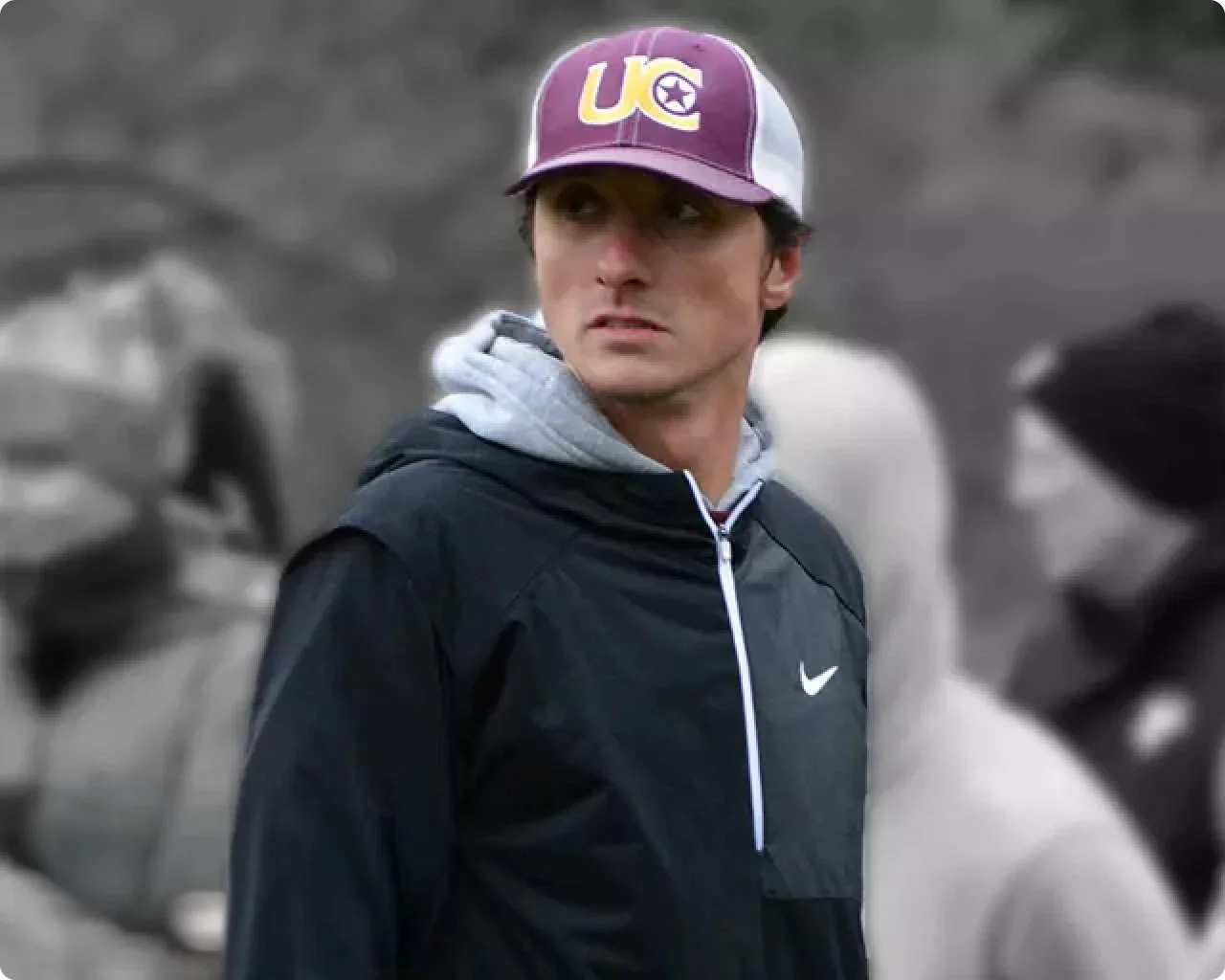 A man wearing a maroon and white baseball cap with the letters "U" and an arrow/star logo, a gray hoodie, and a black Nike jacket, standing outdoors with a blurred background of rocks and other people.