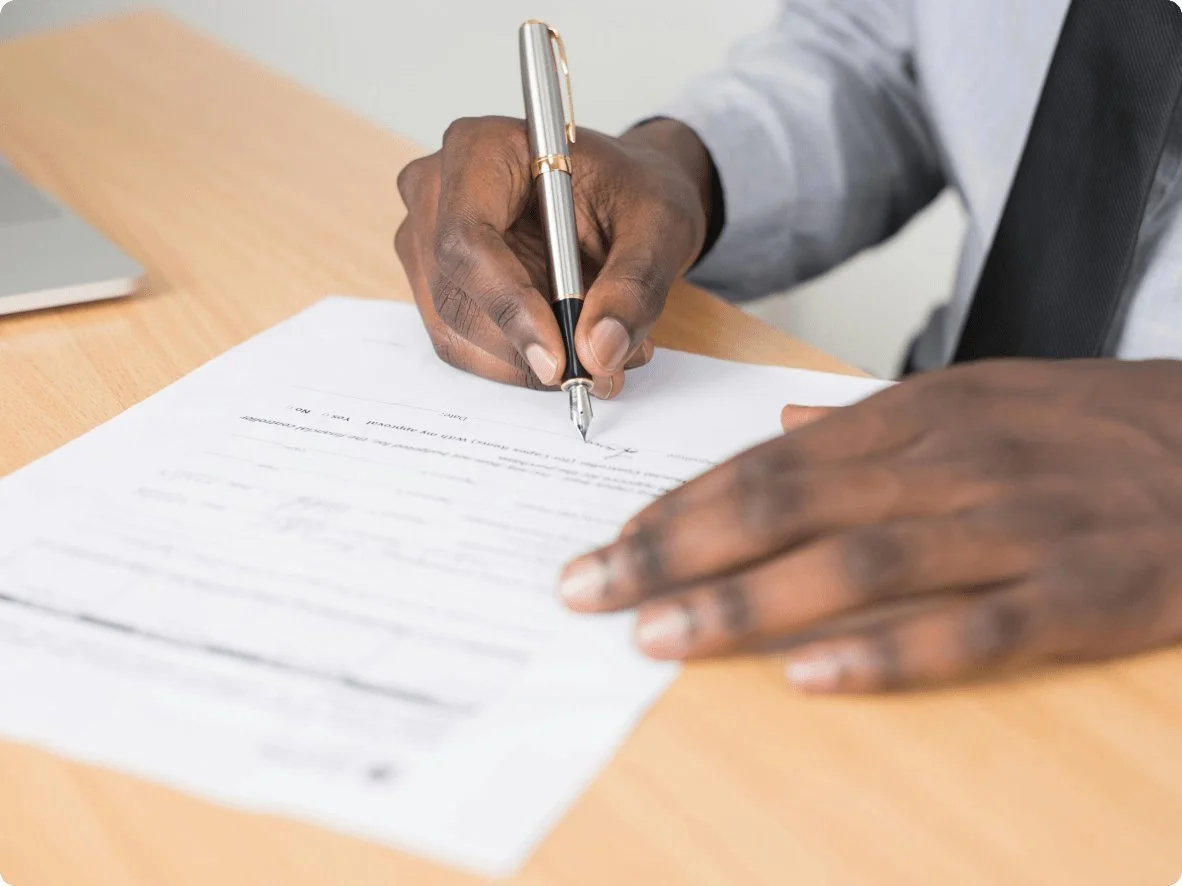 Person signing a document with a pen on a wooden desk.