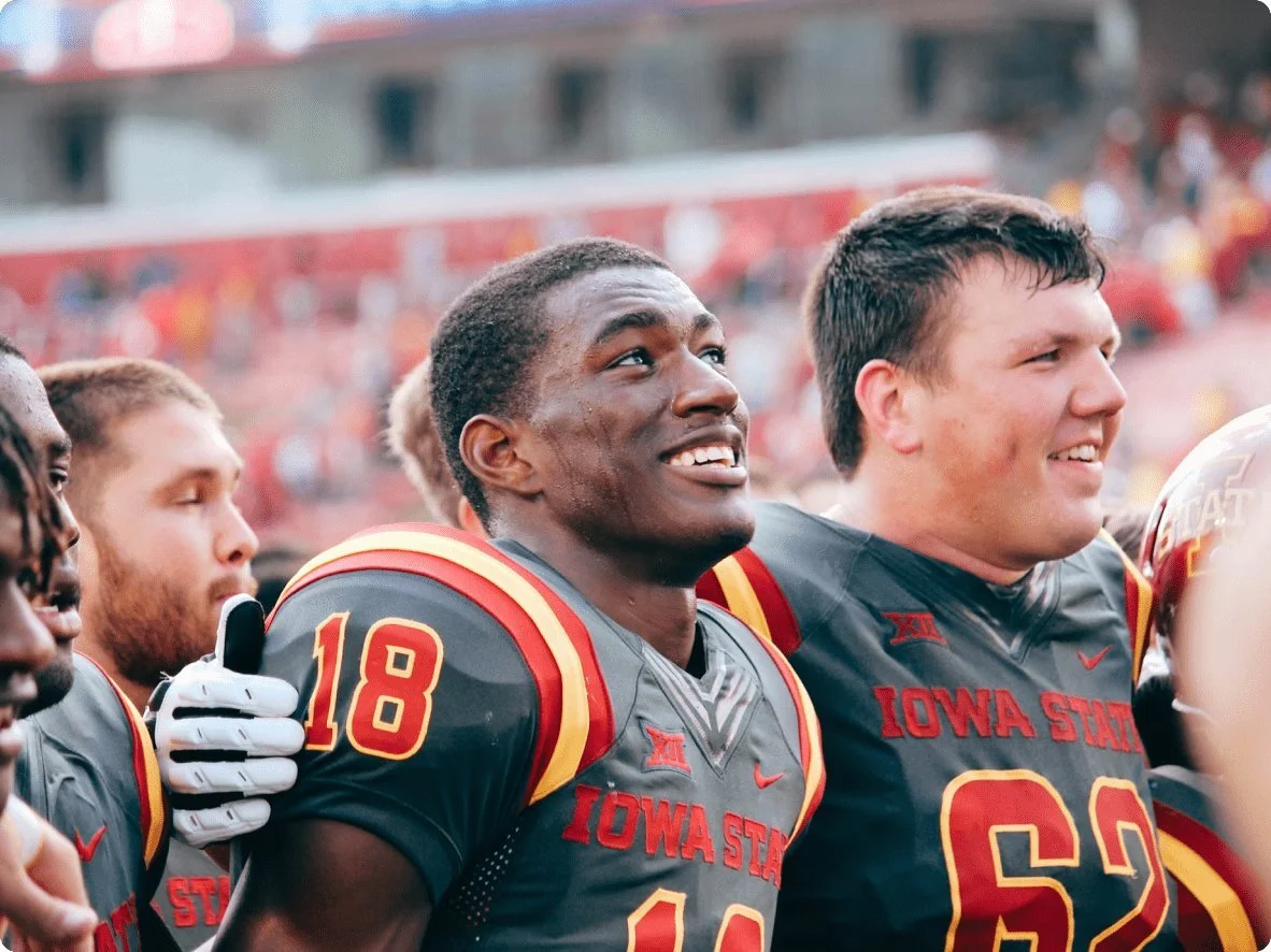 Iowa State football players standing during a game, wearing black jerseys with red and yellow accents, some with eyes closed and others looking ahead.