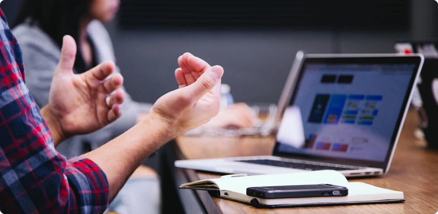 Person in plaid shirt gesturing while discussing in a meeting room with laptops and notebooks on the table.