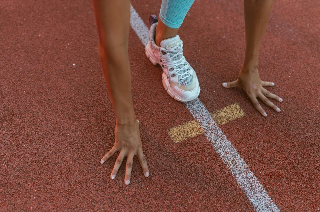 A person at the starting line of a running track in a ready position, wearing white athletic shoes and blue leggings.