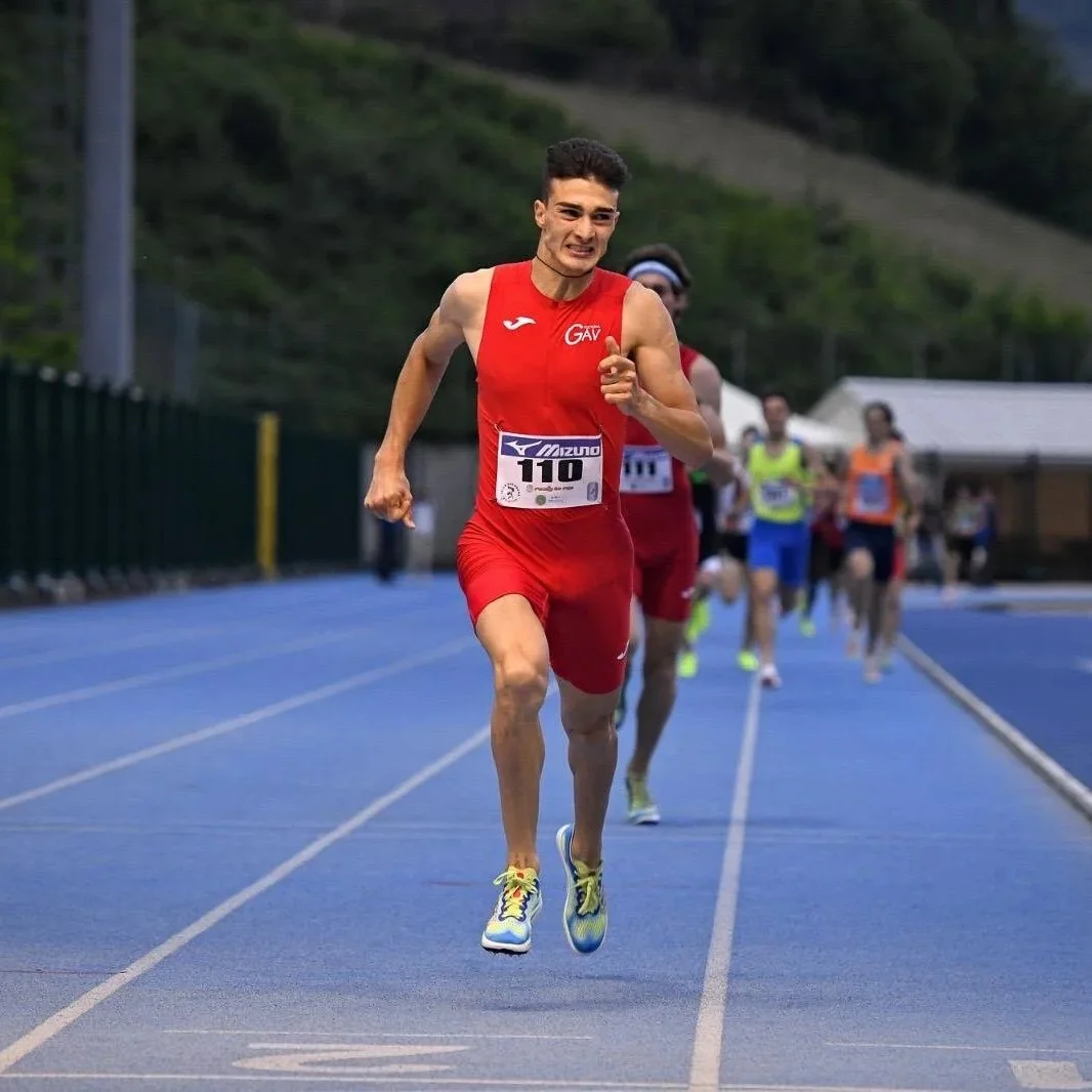 Male athlete in red running uniform with bib number 110 competing in a race on a blue track, with other runners behind him and outdoor setting with trees and tents.