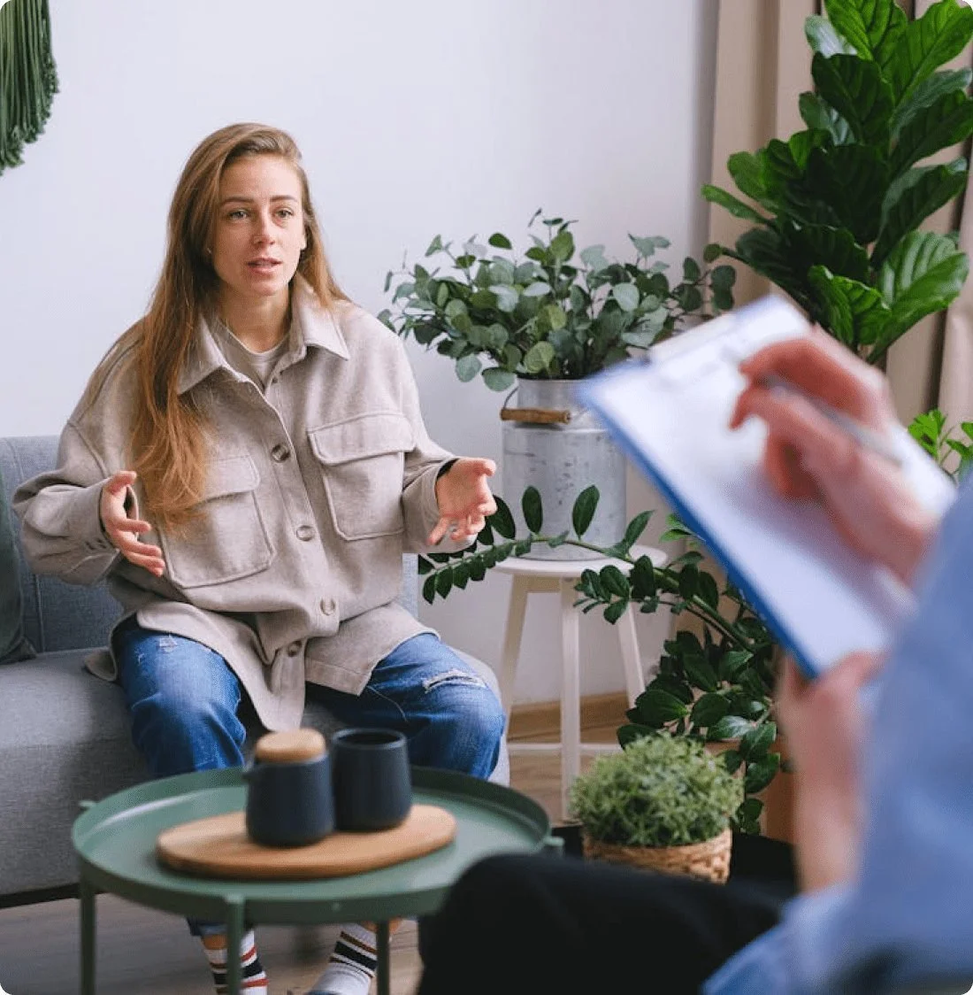 Woman gesturing as if explaining during interview in living room with green plants and coffee table.