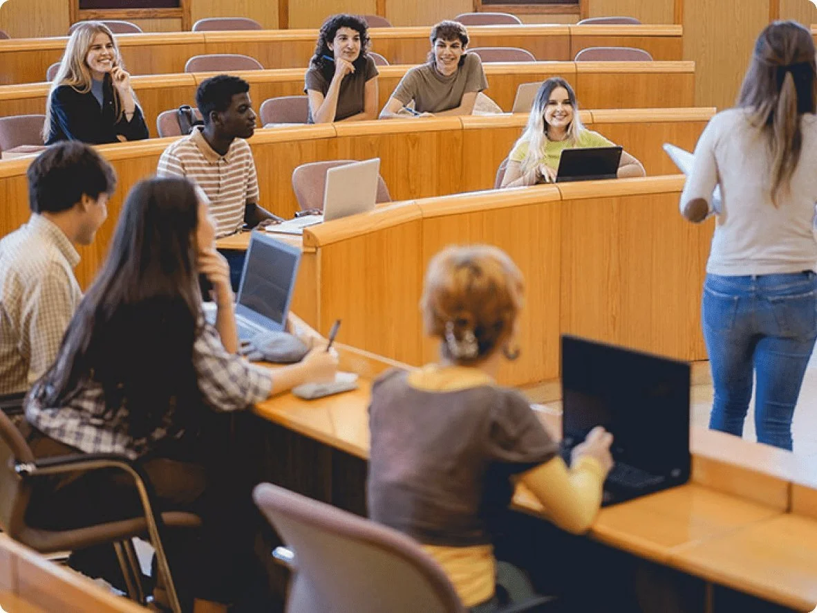 Students in a classroom, some using laptops, listening to a woman standing with a paper, as she teaches or presents to the class.
