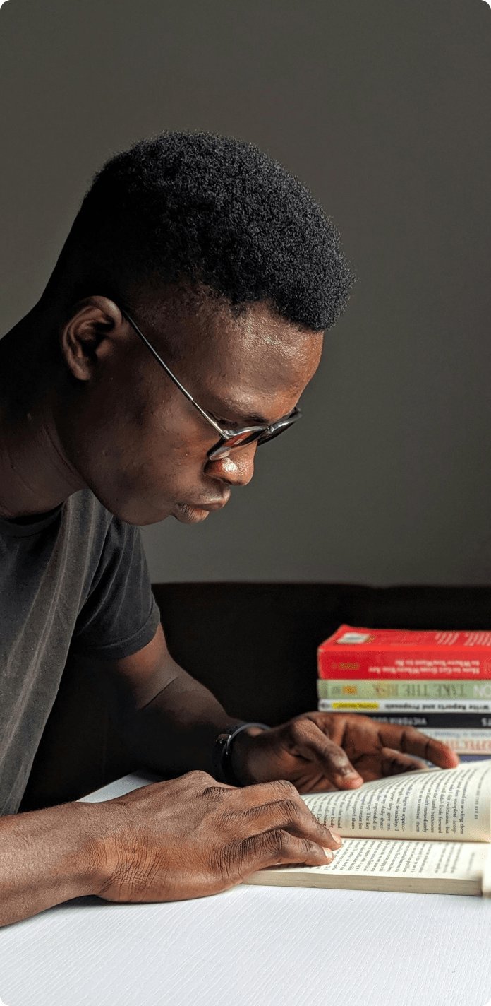 A young man wearing glasses reading a book at a table with several other books stacked nearby.