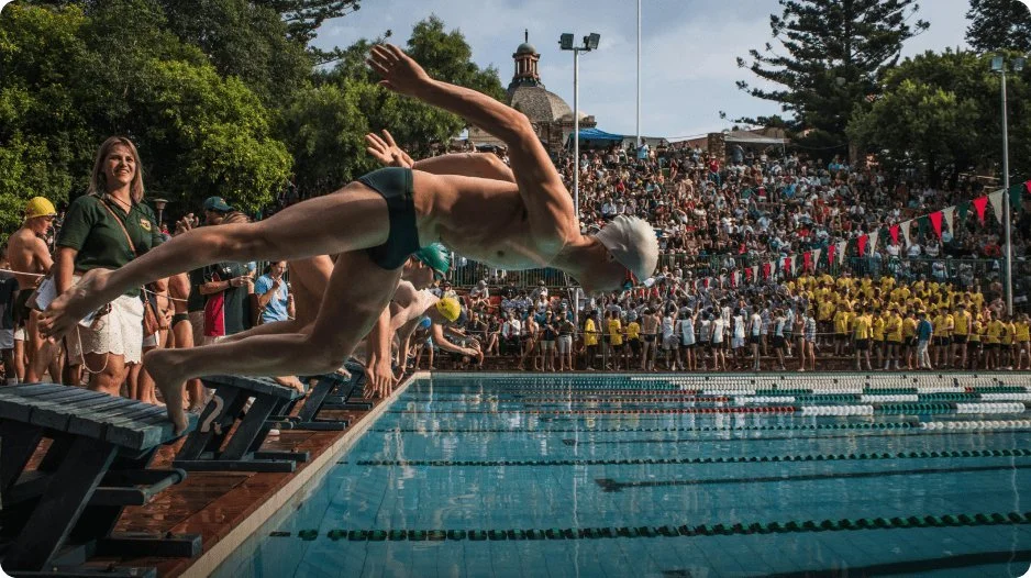 Swimmer dives off starting block into swimming pool during race, with spectators and other swimmers in background.