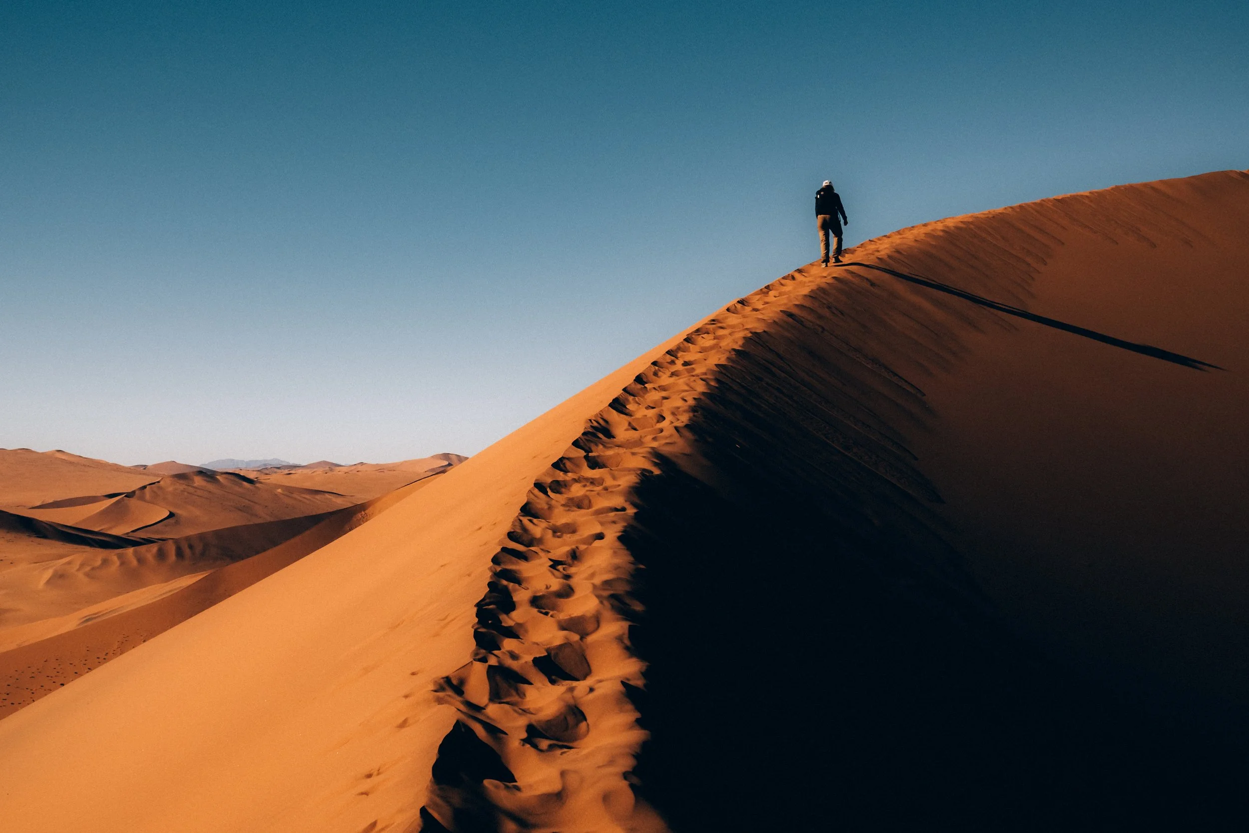 A person in dark clothing and a hat walking along the crest of a sand dune in a desert under a clear blue sky.