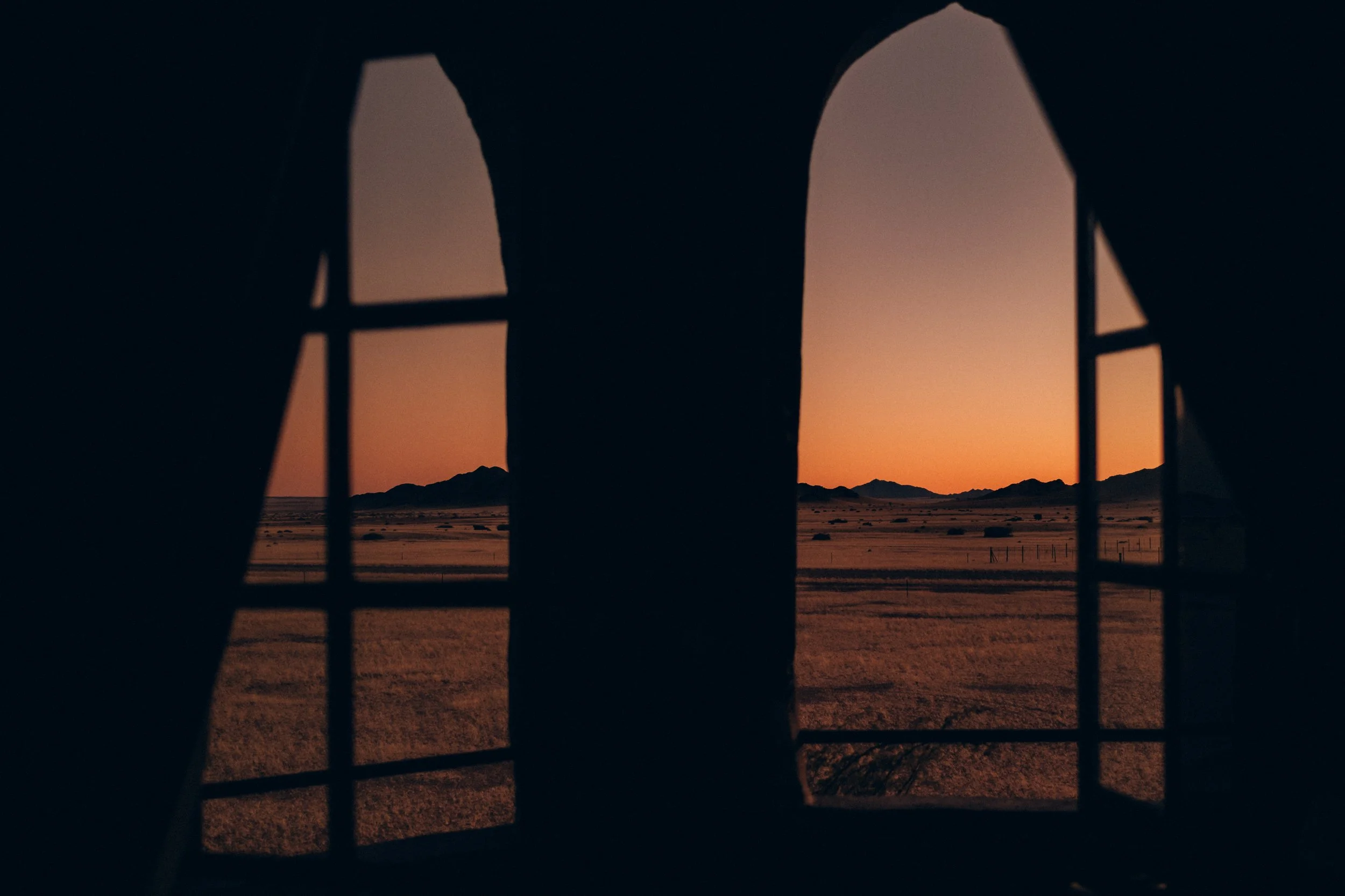 View of a desert landscape at sunset seen through a window with multiple panes and dark curtains.