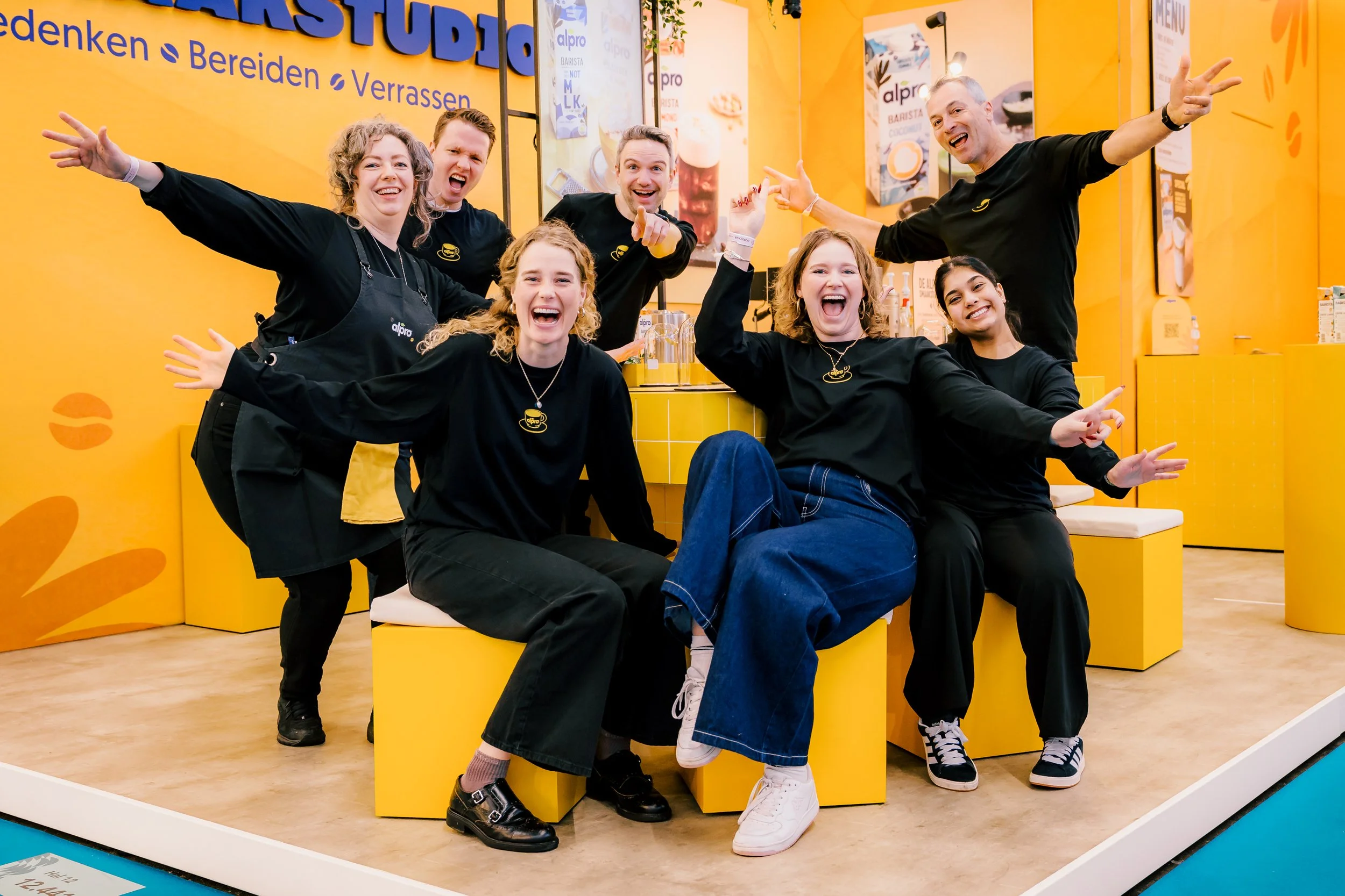 A group of seven people smiling and posing in a bright yellow cafe or stand with orange decor, all wearing black shirts, some with small accessories, enjoying a joyful moment together.