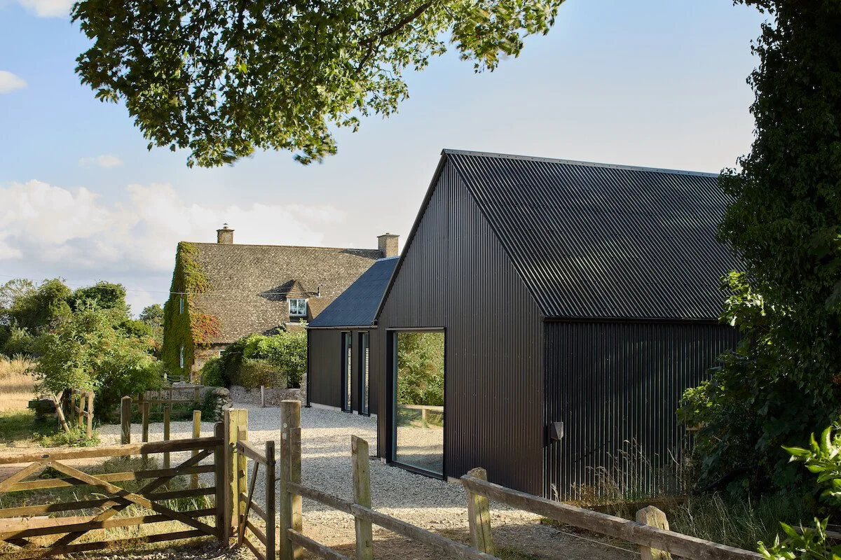 Taw Fitzwilliam Ltd — A modern black metal self-built sustainable house with large windows, set in the Cotswolds with a gravel pathway and surrounded by trees.