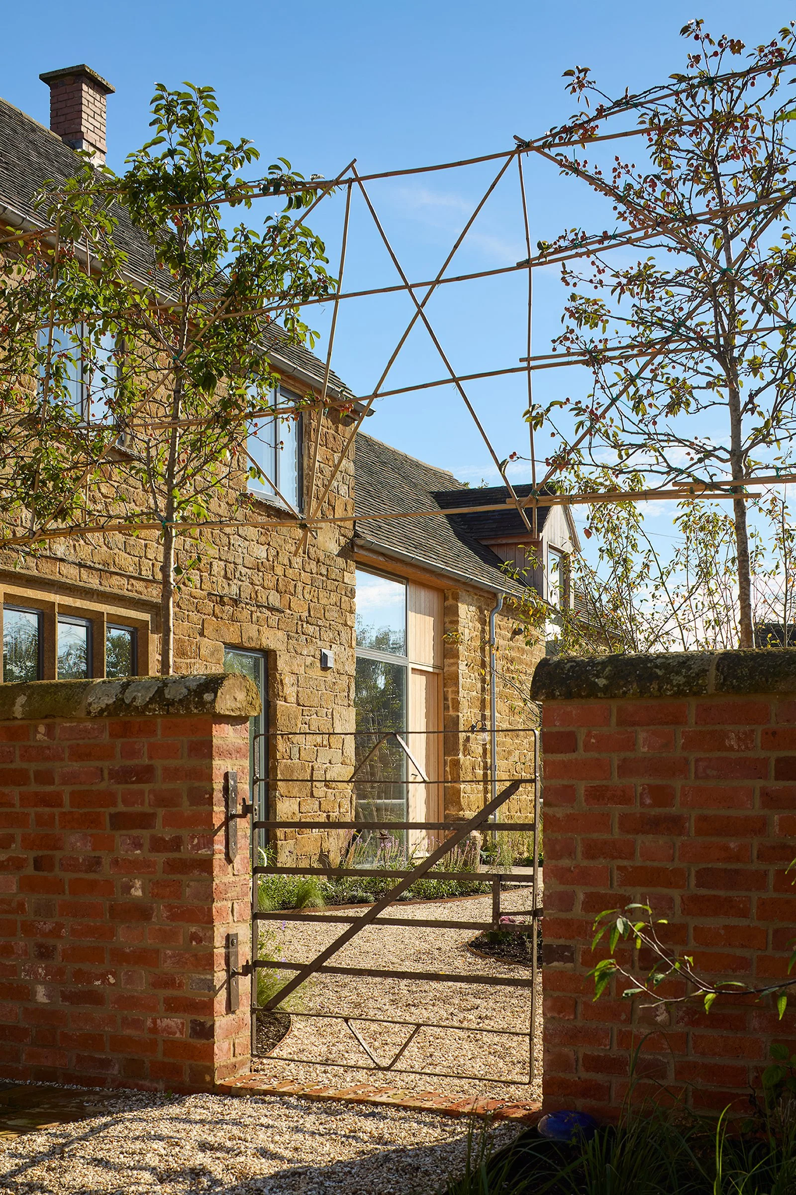 Taw Fitzwilliam Ltd — A renovation of a Grade II listed stone farmhouse on the edge of the Cotswolds, with a tiled roof and large windows, viewed through a metal gate, with