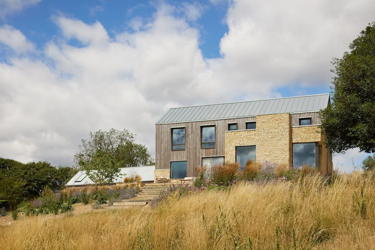 Taw Fitzwilliam Ltd — Contemporary architecturally designed house in the Cotswolds with timber and stone exterior, large windows, metal roof, surrounded by tall grass and t