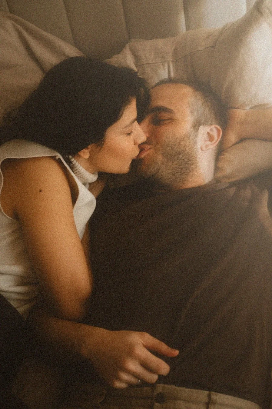 Close-up lifestyle portrait of a couple kissing while lying down on a bed in a Berlin Kreuzberg apartment with warm, grainy textures.