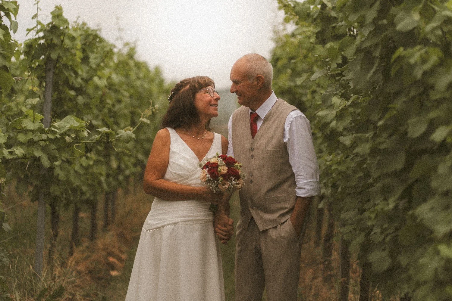 A medium outdoor shot of the wedding couple walking and smiling at each other while holding hands in a lush green vineyard.