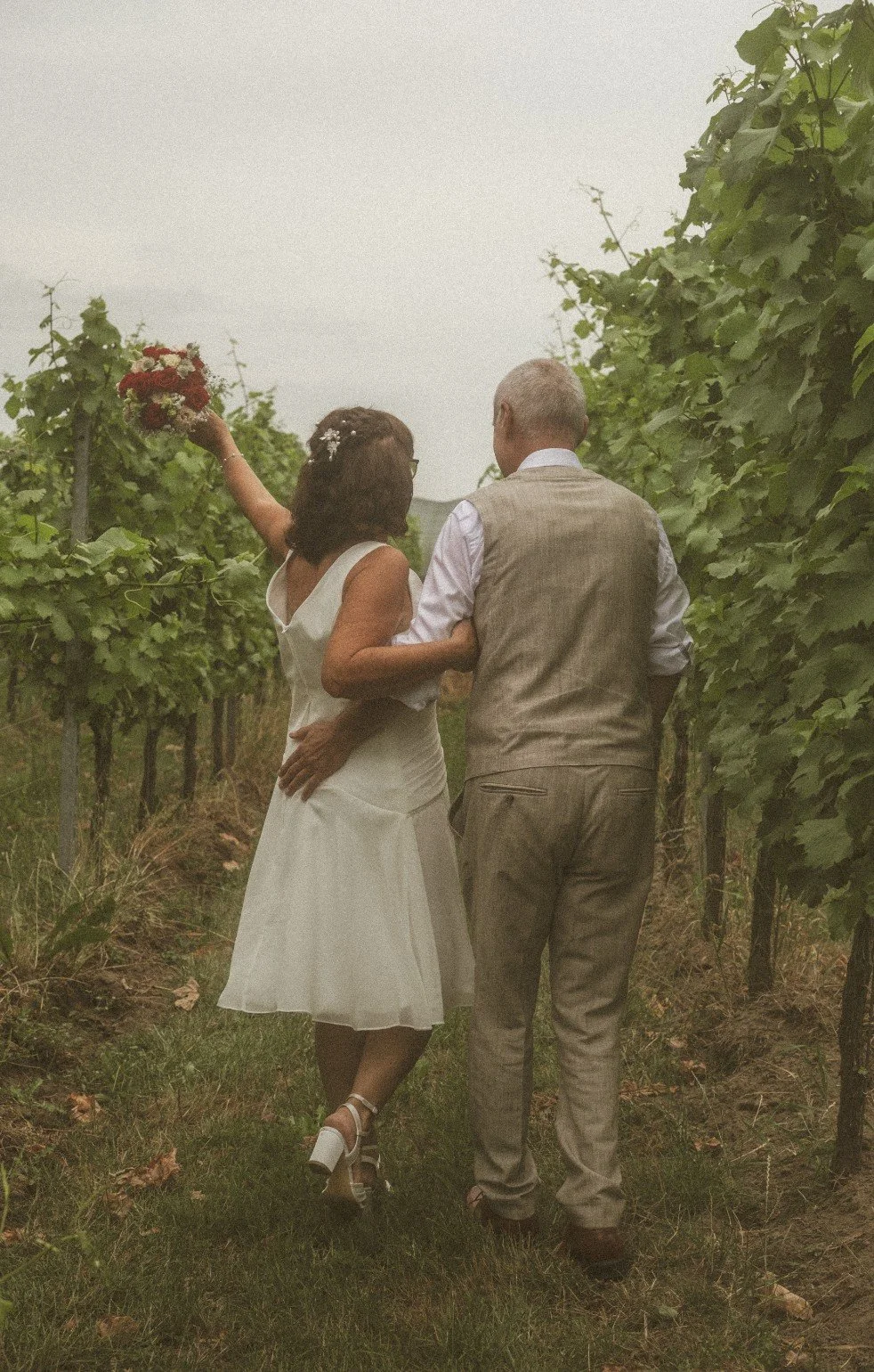 A back view of the bride holding up her bouquet while walking arm-in-arm with the groom through a vineyard.