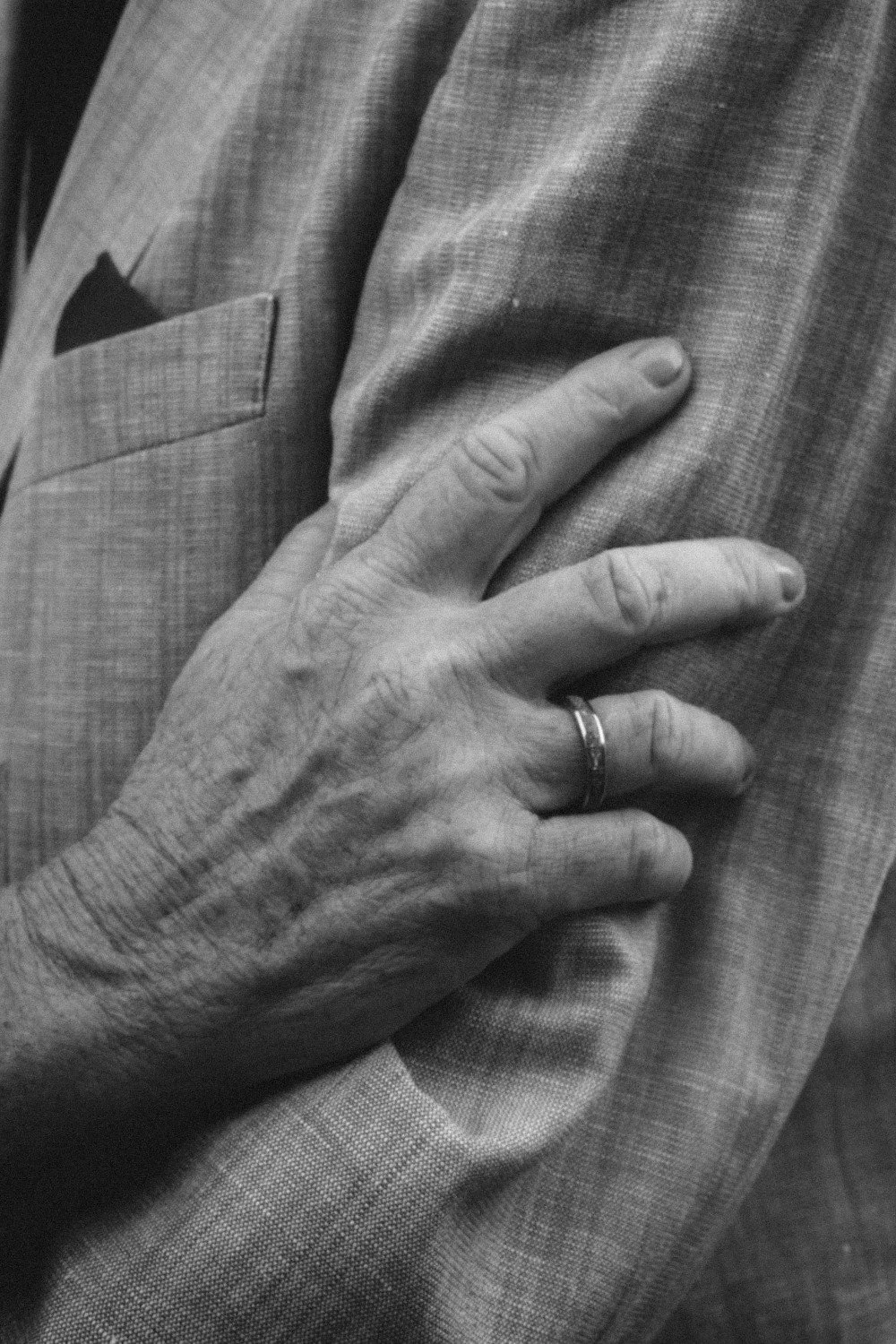 A close-up black and white indoor shot of a hand wearing a wedding band resting gently on a suit jacket sleeve.