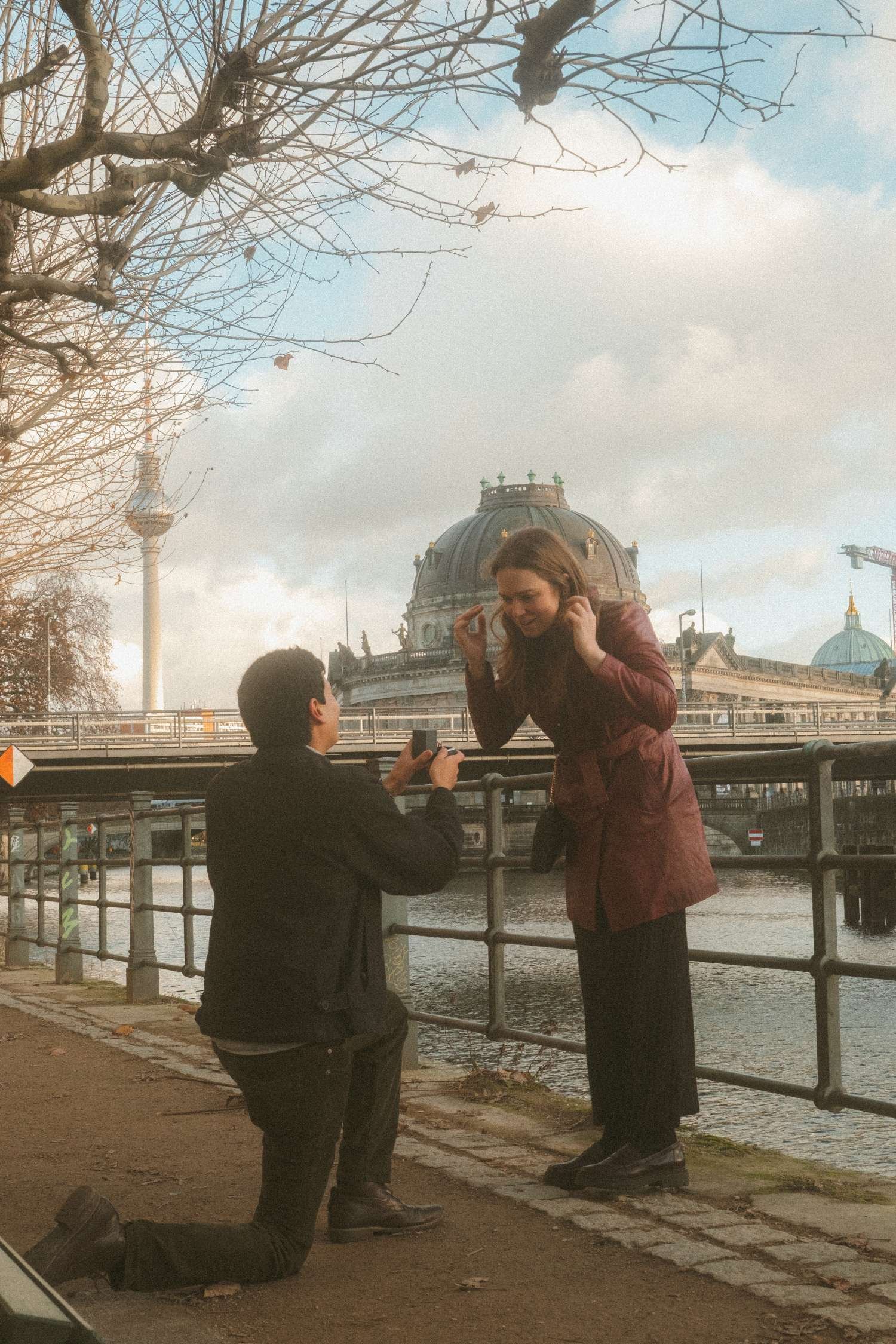 Man kneeling for a surprise proposal by the river as his partner reacts with the Berlin TV Tower and Bode Museum in the background