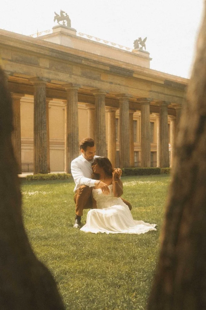 A romantic couple sharing a tender embrace in a grassy park in front of historic neoclassical architecture during an engagement photoshoot in Berlin.